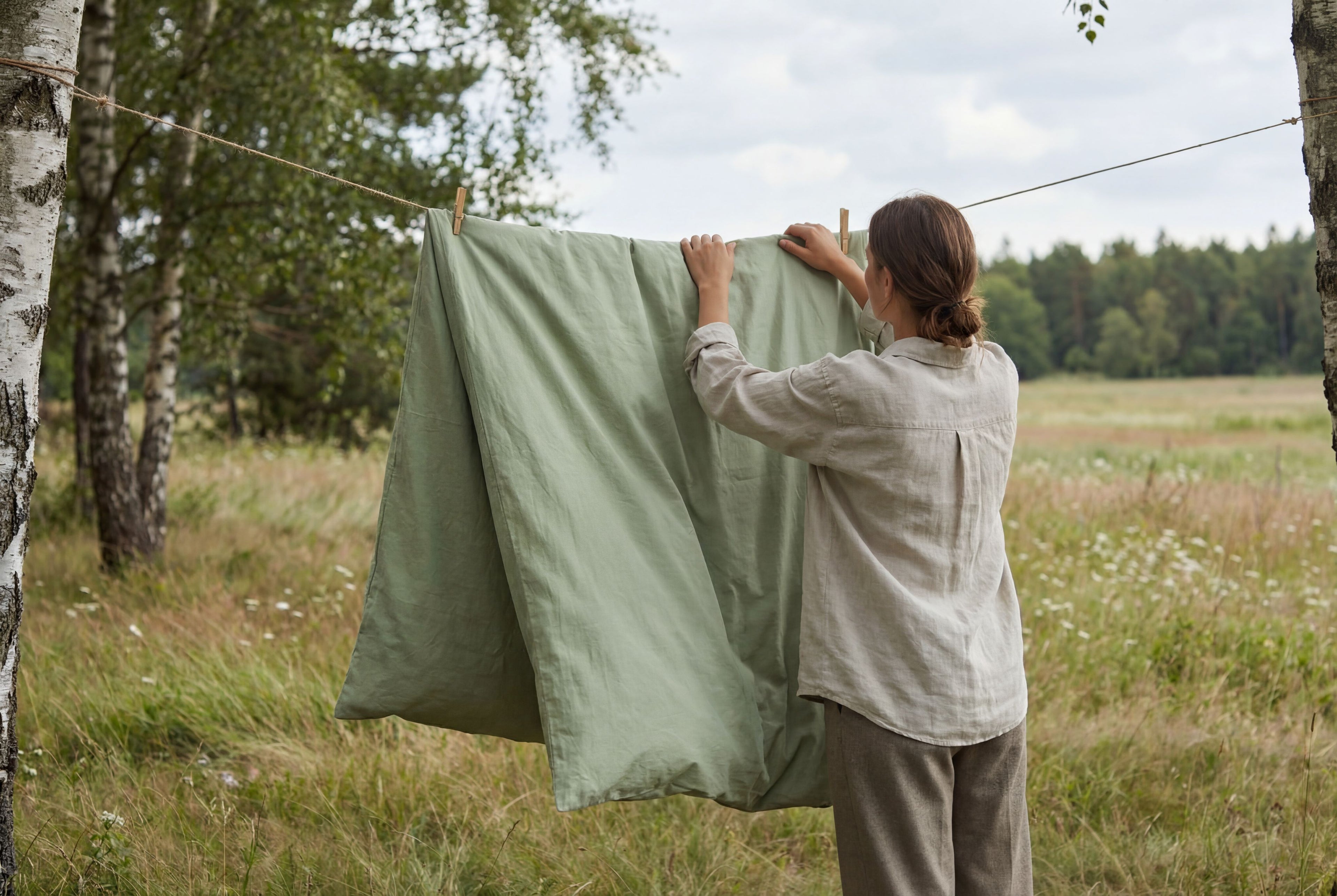 Person hanging a green sheet on a clothesline in a natural setting with trees and grass.