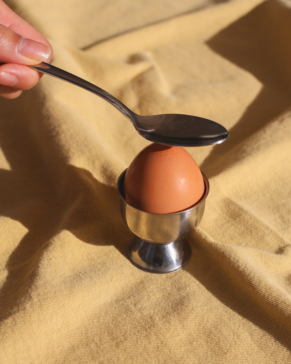 Egg in a cup with a spoon balancing on top, held by a hand against a beige fabric background