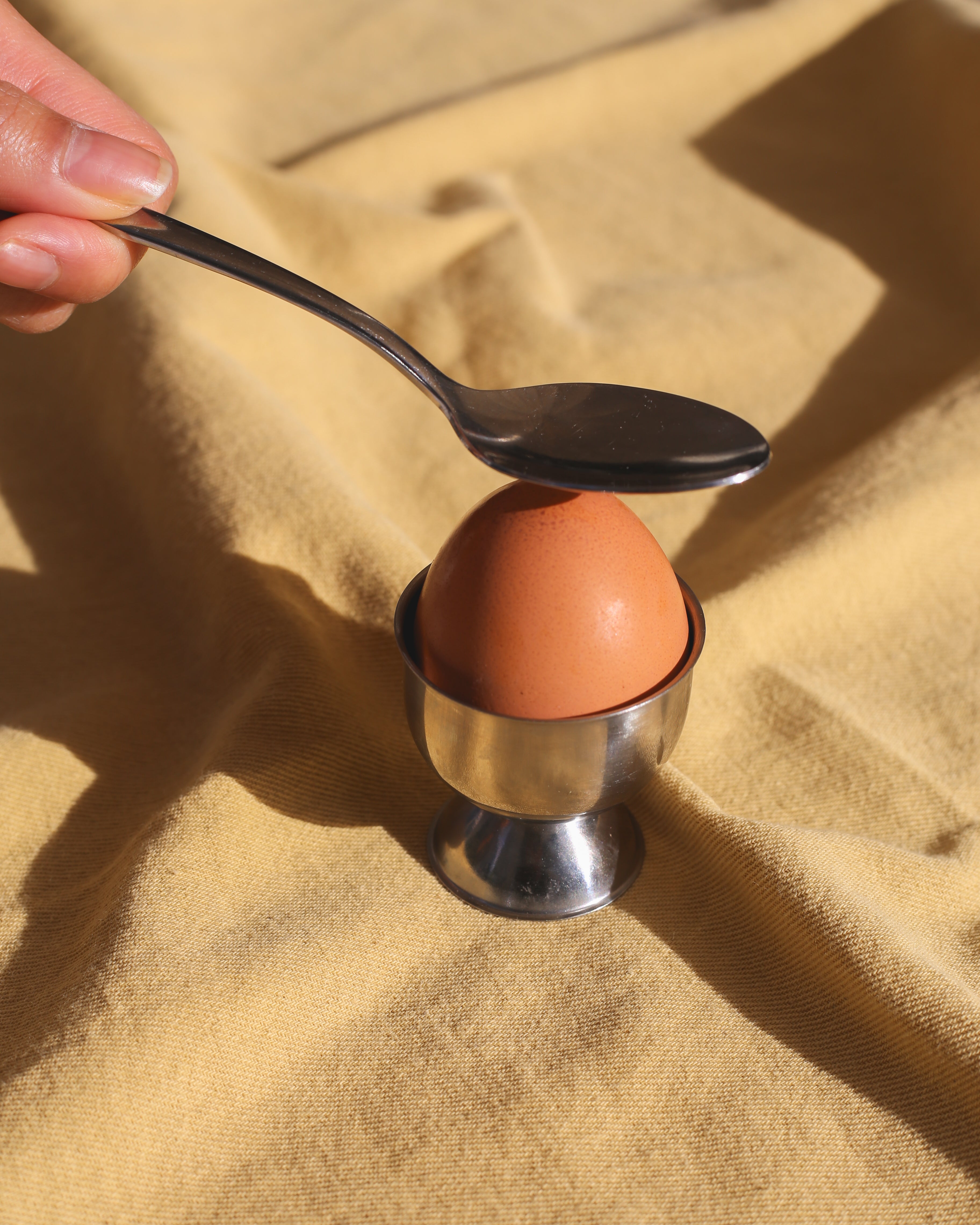 Egg in a cup with a spoon balancing on top, held by a hand against a beige fabric background