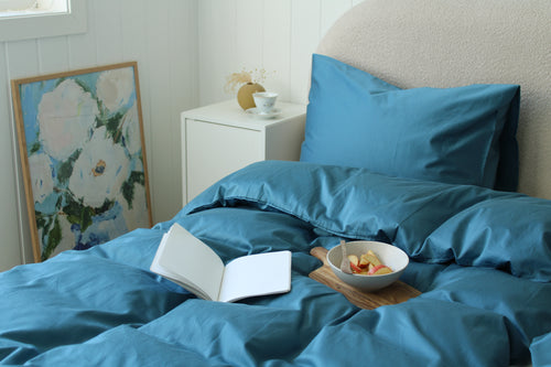Blue bedding set with an open book and fruit bowl on a tray in a bedroom.