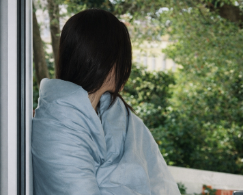 Person wrapped in a light blue blanket on a balcony with greenery in the background