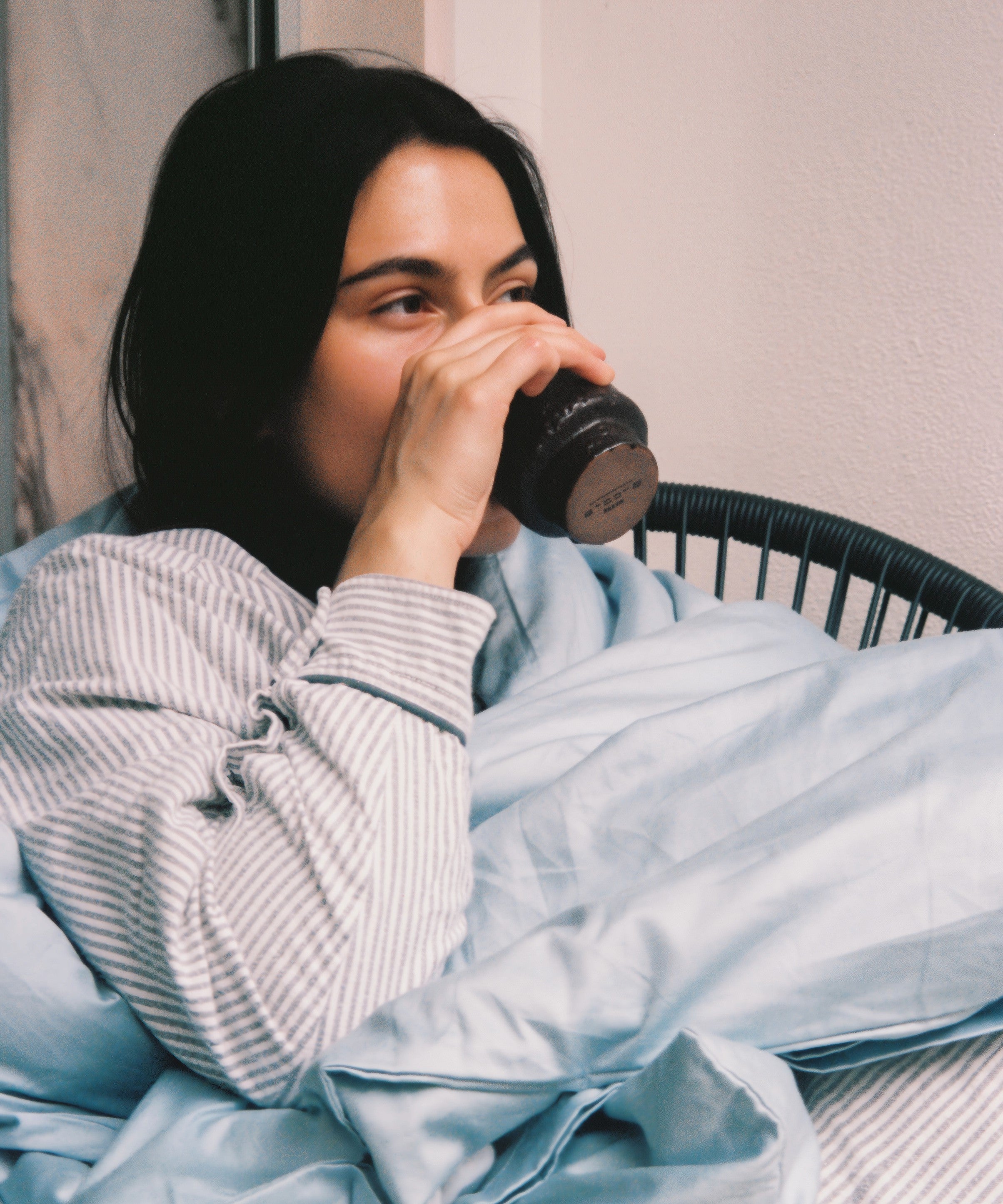 Woman drinking from a mug while wrapped in a blanket on a chair.