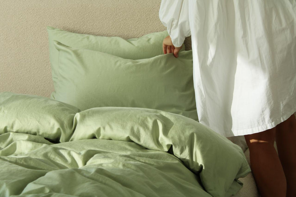 Person in a white robe adjusting green bedding in a bedroom.