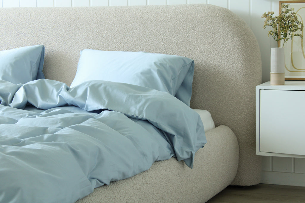 Bed with light blue bedding and pillows against a beige headboard, next to a white nightstand.